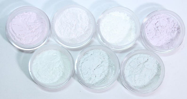 May include: Seven small, clear jars filled with various shades of powder. The colors include white, pale lavender, and several shades of pale green. The powders have a fine, textured appearance, and the jars are arranged in a circular pattern.