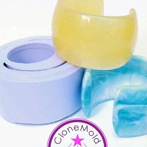 May include: A yellow, translucent cuff bracelet sits atop a light blue bracelet and a purple mold. The cuff is a wide, open design. The image also shows a purple and white logo with the text "CloneMold".