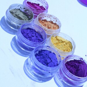May include: An overhead shot of seven small, clear plastic jars filled with colorful powder. The powders are various shades of purple, blue, yellow, orange, and green. The jars are arranged diagonally on a white surface, casting shadows.