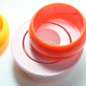 May include: Three plastic bangles stacked on top of each other. The top bangle is orange, the middle bangle is white, and the bottom bangle is pink.