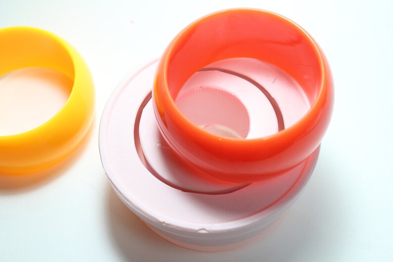 May include: Three plastic bangles stacked on top of each other. The top bangle is orange, the middle bangle is white, and the bottom bangle is pink.