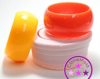 May include: Two silicone moulds for making resin bangles. The moulds are stacked on top of each other. The top mould is orange and the bottom mould is yellow. The moulds are sitting on a white surface. The image also includes a purple circle with the text "Clone Mold" and three pink stars.