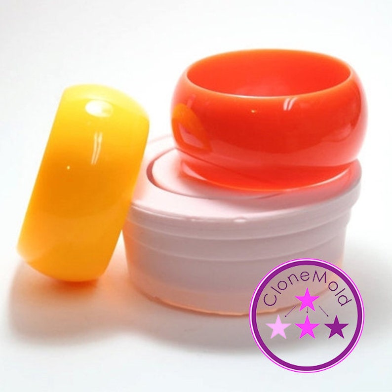 May include: Two silicone moulds for making resin bangles. The moulds are stacked on top of each other. The top mould is orange and the bottom mould is yellow. The moulds are sitting on a white surface. The image also includes a purple circle with the text "Clone Mold" and three pink stars.