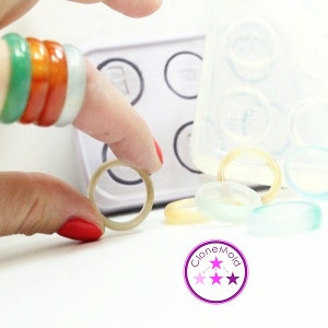 Puede incluir: Una mano sosteniendo un anillo de color claro junto a un molde transparente con varios anillos coloridos. Los anillos son en tonos de verde, naranja y blanco. El logotipo "CloneMold" está en la esquina inferior derecha.