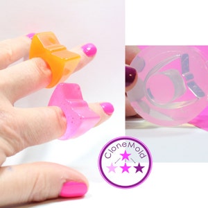 以下が含まれることがあります： 星型の樹脂製のリングが2つ、1つはオレンジ、もう1つはピンク。リングは指にはめられています。画像には、リングの透明なシリコンモールドも写っています。