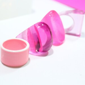 May include: Three pink, clear, glass rings. The rings are different shapes and sizes. The rings are on a white surface.