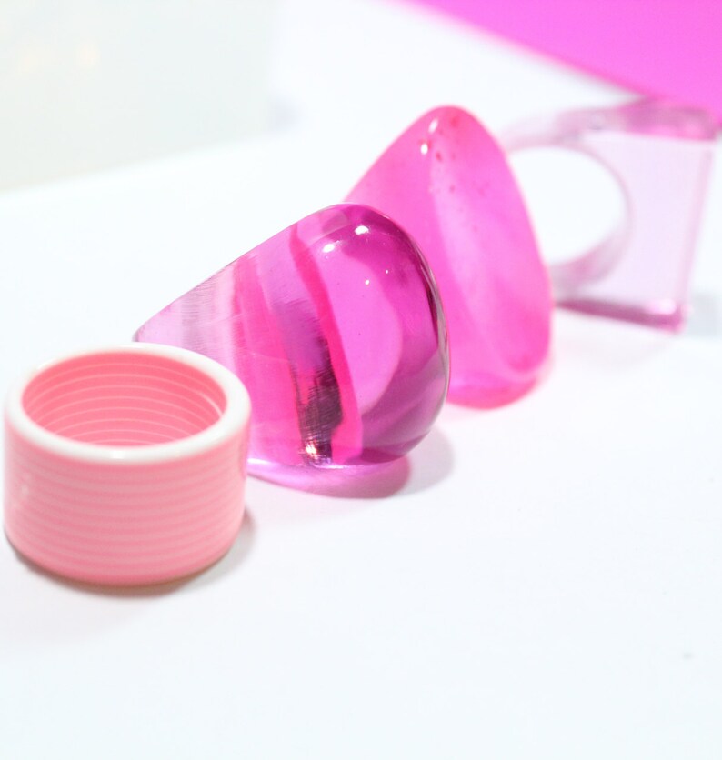 May include: Three pink, clear, glass rings. The rings are different shapes and sizes. The rings are on a white surface.
