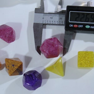 May include: A set of six colorful dice with glitter embedded in the resin. The dice are in various shapes, including a pyramid, a cube, and a dodecahedron. The dice are measured with a caliper, which reads 22.69 mm.