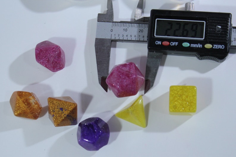 May include: A set of six colorful dice with glitter embedded in the resin. The dice are in various shapes, including a pyramid, a cube, and a dodecahedron. The dice are measured with a caliper, which reads 22.69 mm.