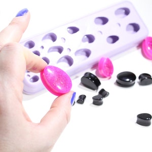 May include: A close-up of a pink glitter ear gauge being held in a hand. The gauge is next to a purple silicone mold with multiple ear gauge shapes. There are also several black ear gauges on a white surface.