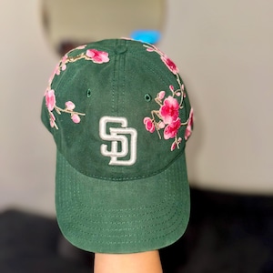 Peut inclure: Casquette de baseball vert foncé avec les lettres "SD" en blanc. La casquette est ornée de broderies florales roses et blanches autour de la visière. Elle possède une visière incurvée et une fermeture à sangle en tissu.