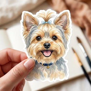 May include: Watercolor sticker of a Yorkshire Terrier dog with a blue bow, collar, and tag. The dog has brown and gray fur, a happy expression, and its tongue is visible. The sticker is held by a hand.