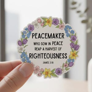 May include: A round sticker with the text "PEACEMAKER" and "WHO SOW IN PEACE REAP A HARVEST OF RIGHTEOUSNESS" surrounded by a floral wreath. The sticker has a white background and is held by a hand.