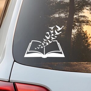 May include: White decal of an open book with birds flying out of it, applied to a car window. The design features a simple, clean aesthetic, suggesting a love for reading and nature. The background shows a sunset.
