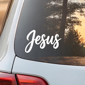 May include: White vinyl decal with the word "Jesus" in a cursive font, affixed to the rear window of a white car. The background shows a blurred view of trees and a sunset.