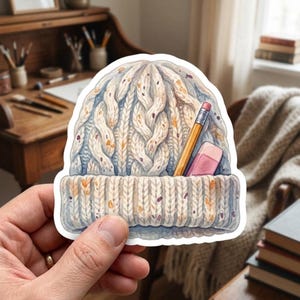 May include: A die-cut sticker featuring a watercolor illustration of a cream-colored cable knit beanie with a folded brim. The beanie has speckled flecks of orange and purple throughout and holds a yellow pencil and a pink eraser in the brim.