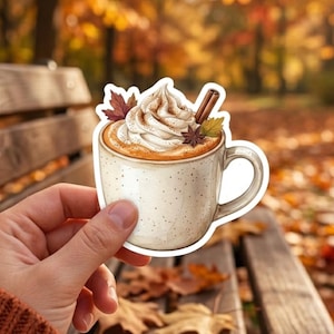 May include: A die-cut sticker of a speckled ceramic mug filled with a warm beverage topped with a swirl of whipped cream, a cinnamon stick, a star anise, and two small autumn leaves in shades of orange, yellow, and brown.