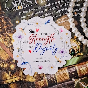 May include: A white sticker with a floral wreath design. The text inside the wreath reads "She is Clothed with Strength and Dignity" with a red heart and a blue heart. The text "Proverbs 31:25" is below the wreath.