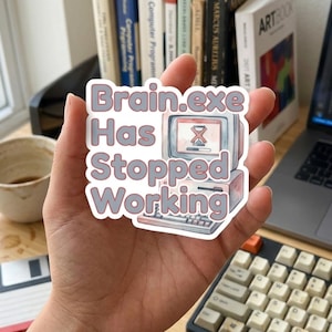 May include: A die-cut sticker featuring a vintage computer illustration with a loading hourglass icon on the screen. The text reads "Brain.exe Has Stopped Working" in bold, light-colored letters with a dark outline, set against a white background.