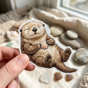 May include: A sticker featuring a watercolor illustration of a smiling otter holding a seashell. The otter has brown fur and is lying on its back. The sticker is held by a hand.