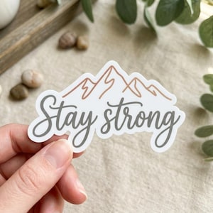 May include: A white die-cut sticker featuring a minimalist line drawing of mountain peaks in a muted terracotta color, positioned above the text "Stay strong" written in a flowing, dark gray cursive script font.