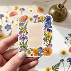 May include: A rectangular sticker frame composed of various pressed flowers and green ferns. The botanical arrangement features vibrant orange poppies, yellow pansies, white daisies, and blue cornflowers, creating a colorful floral border design.