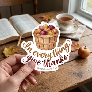 May include: A die-cut sticker featuring a watercolor illustration of a wooden bushel basket filled with red and yellow apples. The text reads In every thing give thanks 1 Thessalonians 5:18 in a brown and maroon script font.
