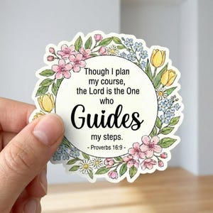 May include: A circular sticker with a floral wreath design. The sticker features the text "Though I plan my course, the Lord is the One who Guides my steps. - Proverbs 16:9 -" in a black script font. The wreath includes pink, yellow, and blue flowers.