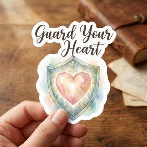 May include: A die-cut sticker featuring a watercolor-style illustration of a pink heart centered within an ornate, light blue shield. Above the shield, the text Guard Your Heart is written in a dark brown, elegant script font against a white background.