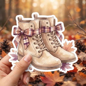 May include: A pair of beige lace-up boots featuring a decorative plaid ribbon tied in a bow around the ankles, accented with illustrated autumn leaves and small berries, presented as a die-cut sticker design with a white border.