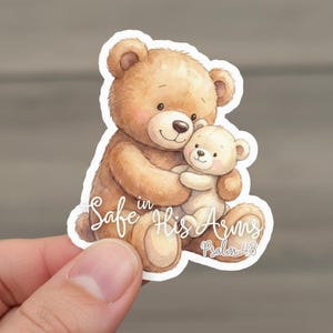May include: A sticker featuring a watercolor illustration of two teddy bears. The larger bear hugs the smaller one. The text "Safe in His Arms Psalm 4:8" is written in cursive. The bears are shades of brown and beige.