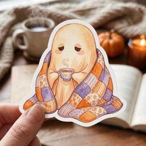 May include: A die-cut sticker featuring a pale, beige ghost with a sleepy expression wrapped in a patchwork quilt with orange, purple, and floral patterns. The ghost holds a small, steaming teacup with both hands, creating a cozy scene.