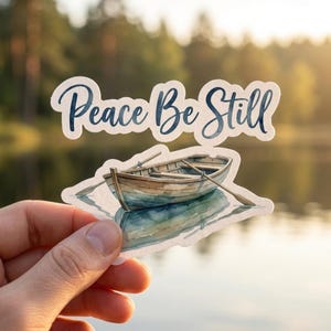 May include: A sticker featuring a watercolor illustration of a rowboat with oars, reflecting in the water. The sticker has the text "Peace Be Still" in a dark blue script font. The background is a blurred image of a lake and trees.