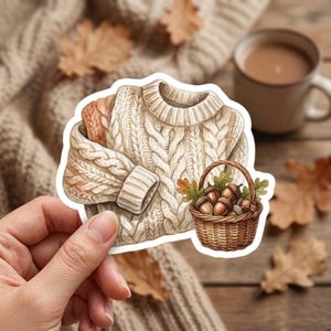 May include: A die-cut sticker featuring a cream-colored cable knit sweater with a ribbed collar and cuffs, paired with a small brown woven basket filled with several brown acorns and green oak leaves, all rendered in a detailed illustrative style.