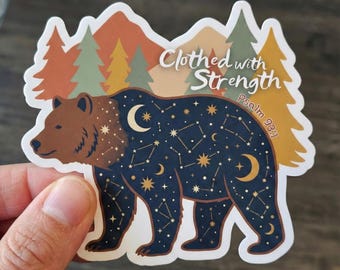 May include: A sticker featuring a brown bear with a constellation design on its body, set against a backdrop of mountains and trees. The text "Clothed with Strength" and "Psalm 93:1" are displayed on the sticker.