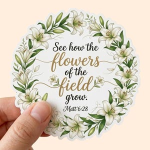May include: A round sticker featuring a floral wreath of white lilies and green leaves. The sticker has the text "See how the flowers of the field grow. Matt 6:28" in black and gold script.