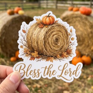 May include: A die-cut sticker featuring a round hay bale topped with a small orange pumpkin, surrounded by scattered autumn leaves and bare twigs, with the gold cursive text Bless the Lord printed at the bottom of the design.