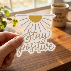 May include: A die-cut sticker featuring a mustard yellow semi-circle sun with radiating lines above the cursive text "Stay positive" in a light brown color. The sticker has a white border and is shaped to follow the outline of the design.