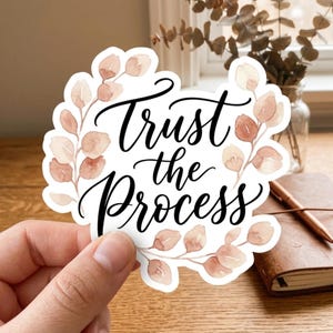 May include: A die-cut sticker featuring the black cursive text Trust the Process centered within a frame of watercolor-style peach and light brown eucalyptus leaves. The sticker has a white border and a matte finish, showcasing delicate botanical art.