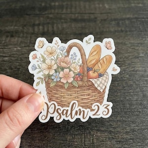 May include: A sticker featuring a woven basket filled with flowers, bread, and fruit. The basket has a handle and is lined with a checkered cloth. The text "Psalm 23" is written in a script font below the basket.