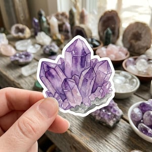 May include: A watercolor sticker of a cluster of amethyst crystals. The sticker features shades of purple and gray, with a white border. The crystals are arranged in a natural, clustered formation, and the sticker is held in a hand.