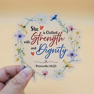 May include: A clear sticker with a floral wreath design. The sticker features the text "She is Clothed with Strength and Dignity" in red and blue script. The Bible verse Proverbs 31:25 is also included.
