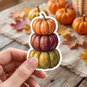 May include: A die-cut sticker featuring a vertical stack of three watercolor-style pumpkins. The top pumpkin is orange, the middle is a deep burgundy, and the bottom is a muted olive green. Each pumpkin has a distinct stem and ribbed texture.