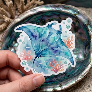 May include: A watercolor sticker featuring a manta ray in shades of blue and purple, with coral and small fish. The sticker has a white border and is held against a background of a seashell.