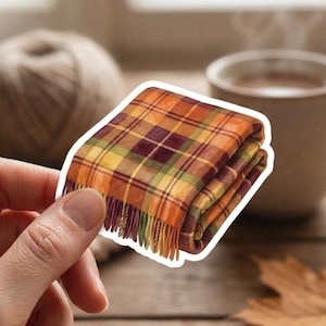 May include: A die-cut sticker featuring a folded throw blanket with a plaid pattern in shades of orange, yellow, burgundy, and green. The blanket has a fringed edge at the bottom and is rendered in a realistic style with a white border outline.