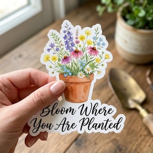 May include: A die-cut sticker featuring a terracotta-colored flower pot filled with a colorful bouquet of purple, yellow, pink, and white wildflowers. Below the pot, the text reads "Bloom Where You Are Planted" in a black cursive script font.