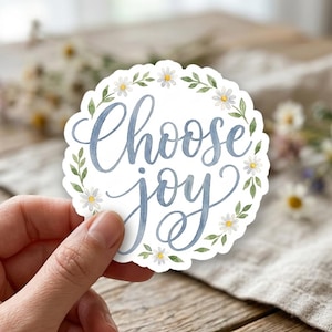 May include: A circular sticker with a scalloped edge featuring the blue cursive text Choose joy in the center. The text is surrounded by a watercolor wreath of small white daisy flowers with yellow centers and green leaves on a white background.