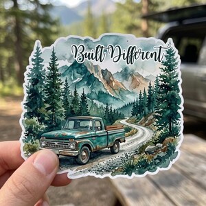 May include: A die-cut sticker featuring a watercolor illustration of a teal vintage pickup truck on a winding dirt road. The scene includes tall green pine trees and jagged mountains in the background. The text Built Different is printed in black script at the top.