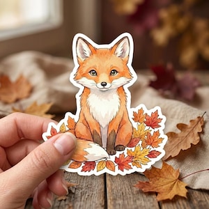 May include: A sticker featuring a cartoon fox with orange fur, a white chest, and a fluffy tail. The fox is sitting among colorful autumn leaves. The sticker is held by a hand on a wooden surface.