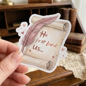 May include: A sticker featuring a scroll with the text "He first Loved Us..." and a feather. The scroll is beige with brown accents, and the feather is pink and brown. Small pink hearts are scattered around the scroll.